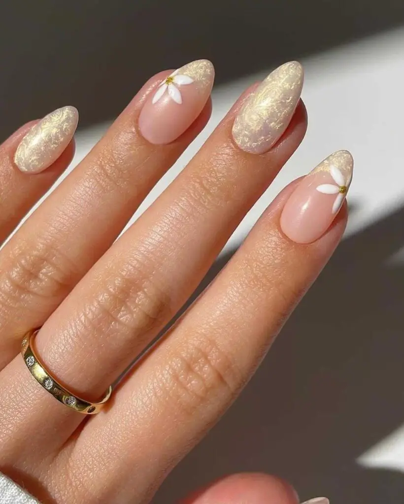A hand with a sheer pink manicure adorned with golden foil and white floral appliqués, embodying understated elegance.