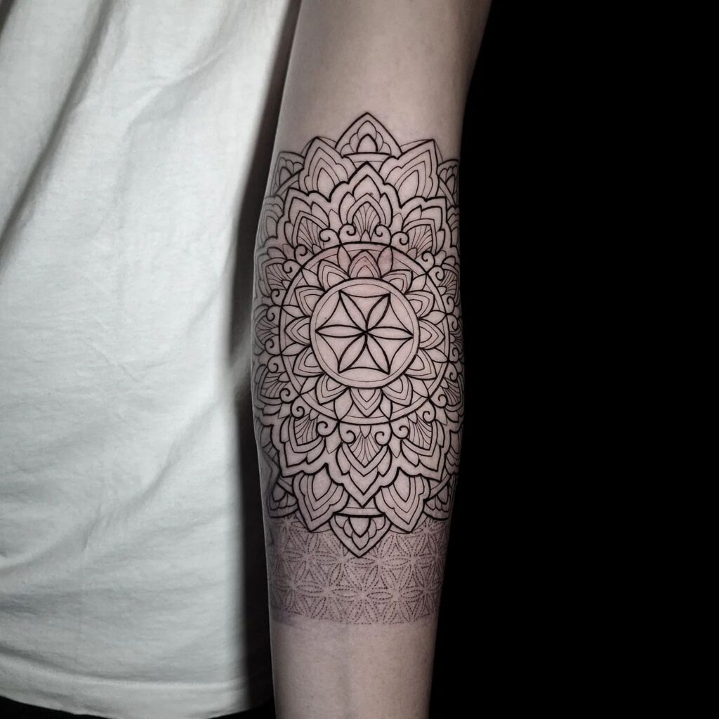 A detailed black ink tattoo on a person's forearm featuring a lotus mandala with a central star-like pattern.