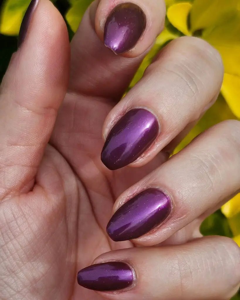 A close-up of a hand with almond-shaped nails polished in a metallic plum shade, set against a springtime backdrop of yellow flowers, showcasing a trendy spring nail color.