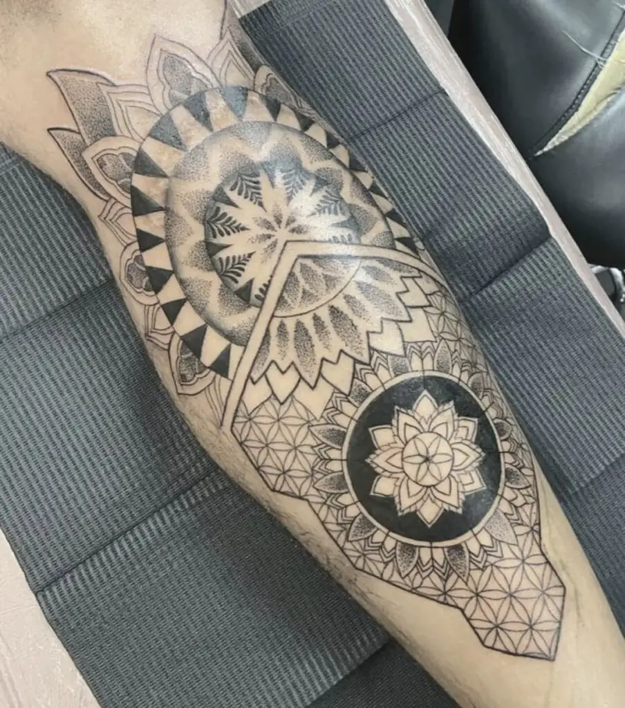 A forearm tattoo depicting a circular mandala with a shield-like appearance, featuring angular and curved patterns.