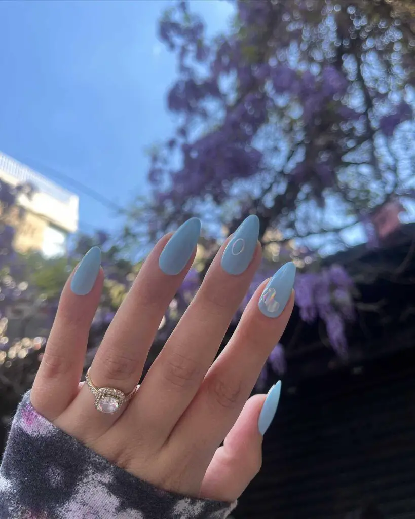 A hand with sky blue almond-shaped nails, featuring subtle white accents, harmonizing beautifully with the purple wisteria backdrop.