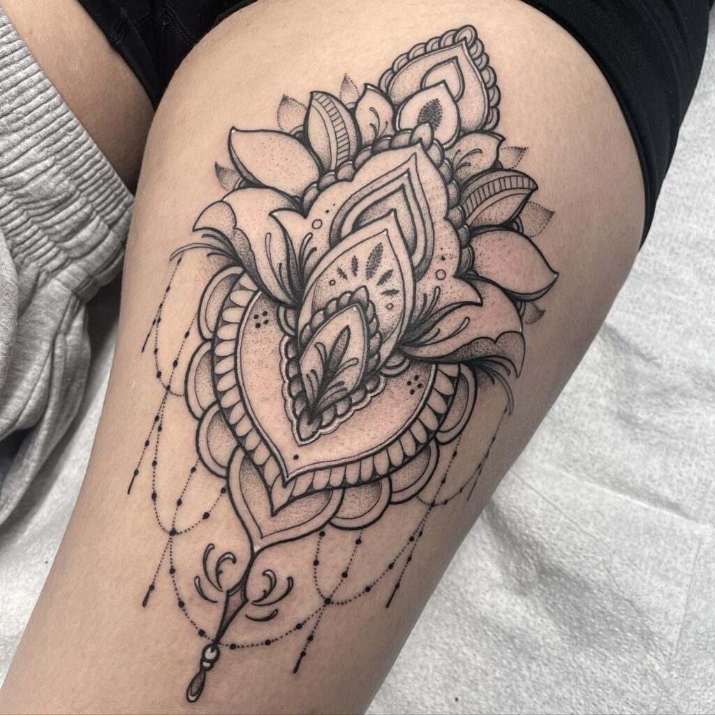 An upper thigh tattoo blending baroque ornamental details with a heart-shaped mandala design.