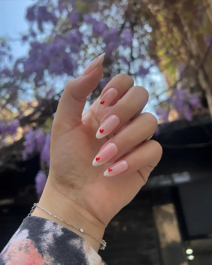 Hand with classic French tip nails, each adorned with a small red heart, set against a wisteria background for a touch of romance.
