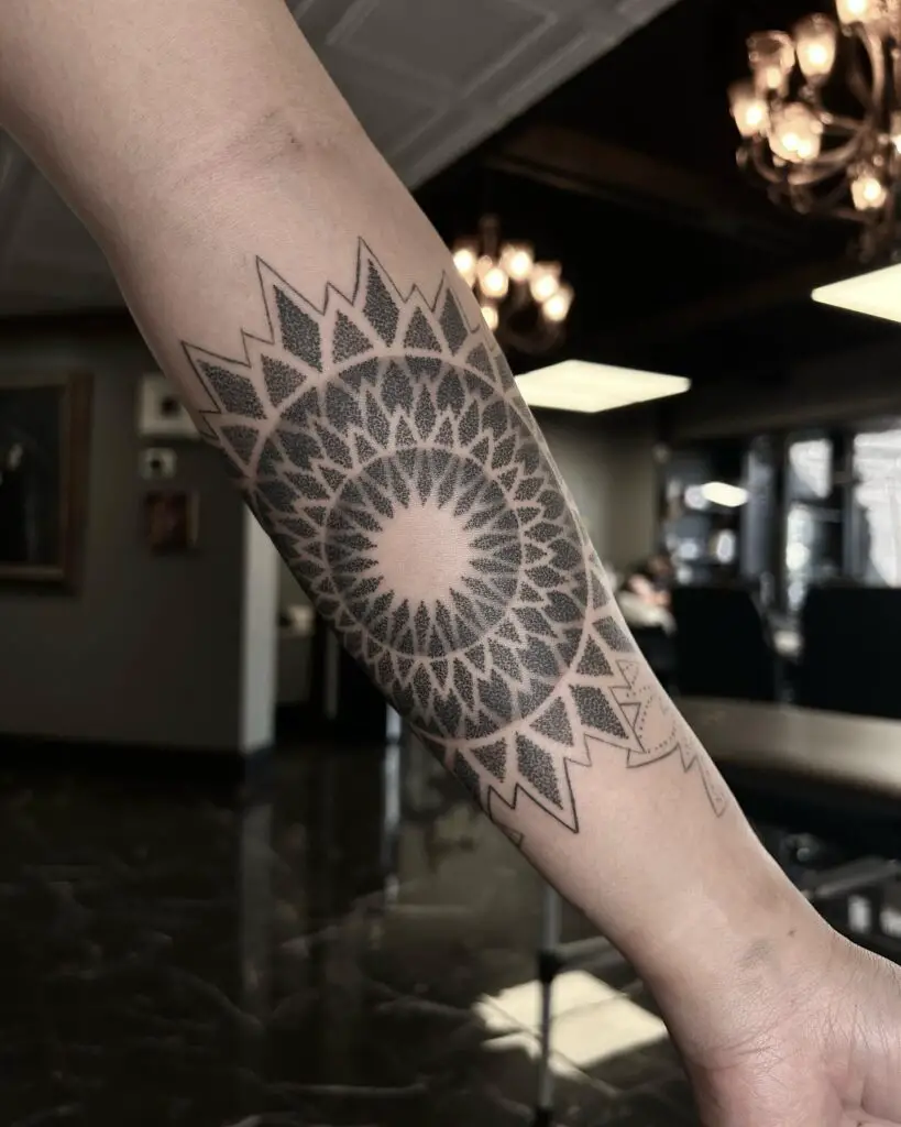 A black ink tattoo on the forearm with a radiant, sun-like mandala center and tribal-inspired edges.