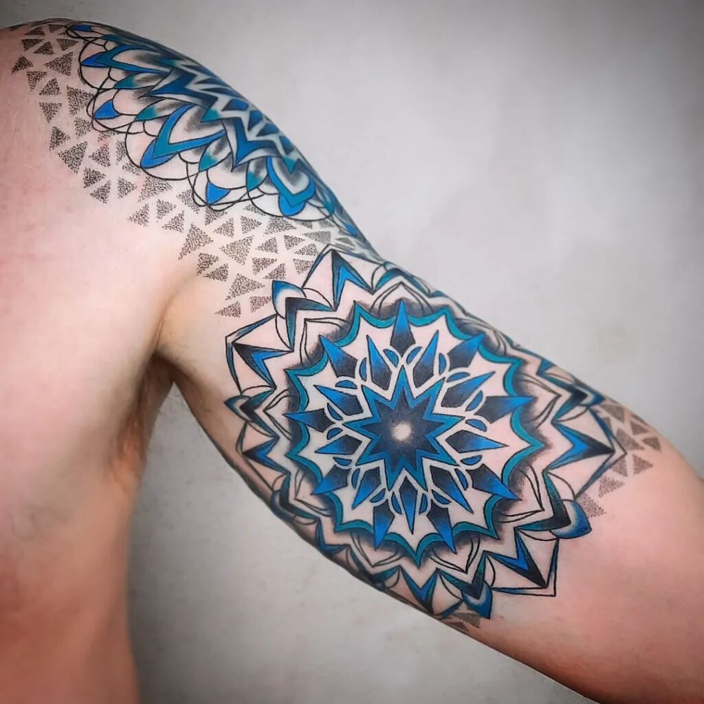 An upper arm tattoo with a central mandala highlighted in shades of blue, creating a three-dimensional effect.