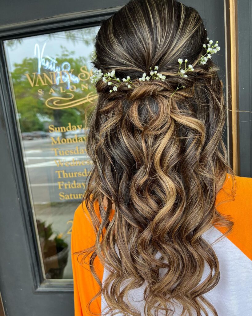 A half-up hairstyle with twisted sections and white floral accents, combined with sun-kissed, wavy curls for a charming prom or spring event look.