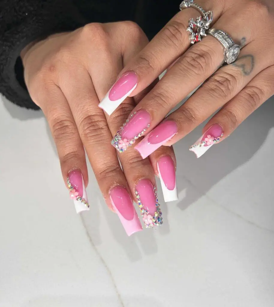 Long nails with a pink base and sparkling French tips, adorned with floral details and rhinestones for an elegant touch.