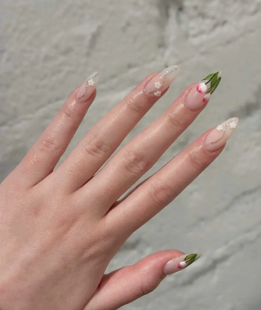 Stiletto nails with a spring-themed design including white floral patterns, a green leaf motif, and delicate glitter accents.