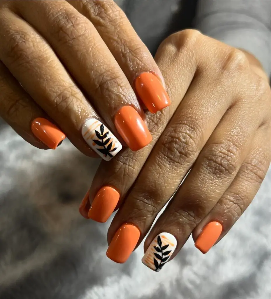 Hands with bright tangerine nail polish, with two accent nails showcasing intricate black botanical leaf designs on a white base.