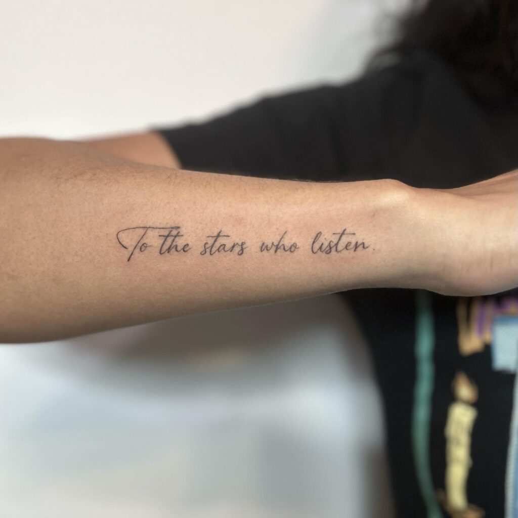 Cursive forearm tattoo saying "To the stars who listen," symbolizing cosmic wonder and poetic reflection.