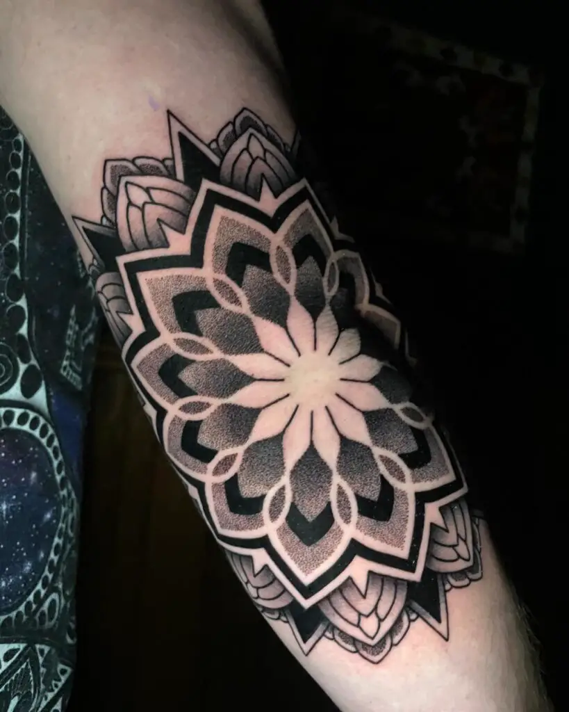 A bold black and shaded mandala tattoo on the shoulder, featuring prominent petal designs and contrasting depths.