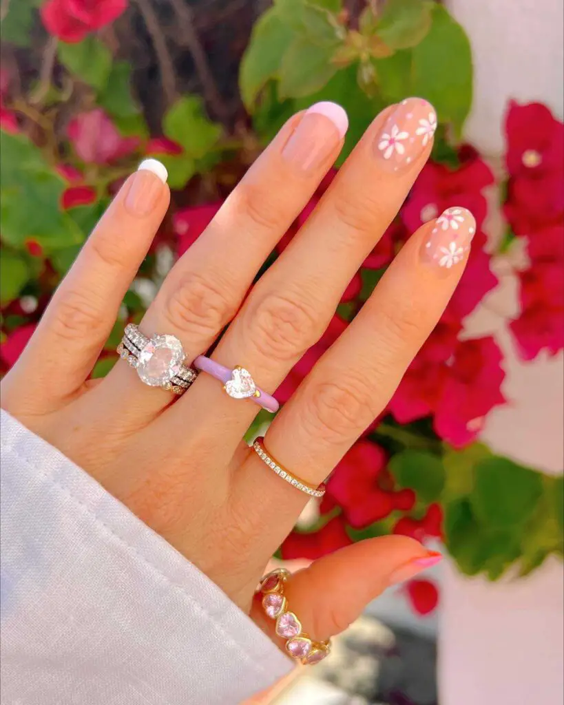 A hand with floral French manicure design against a backdrop of pink blossoms, showcasing white tips with flower accents on sheer pink base.