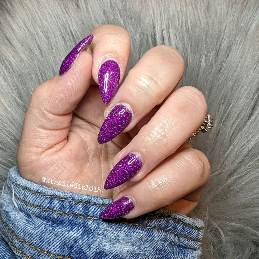 Hand draped in blue fabric, displaying long nails covered in bold purple glitter, conveying a sense of luxury and confidence.